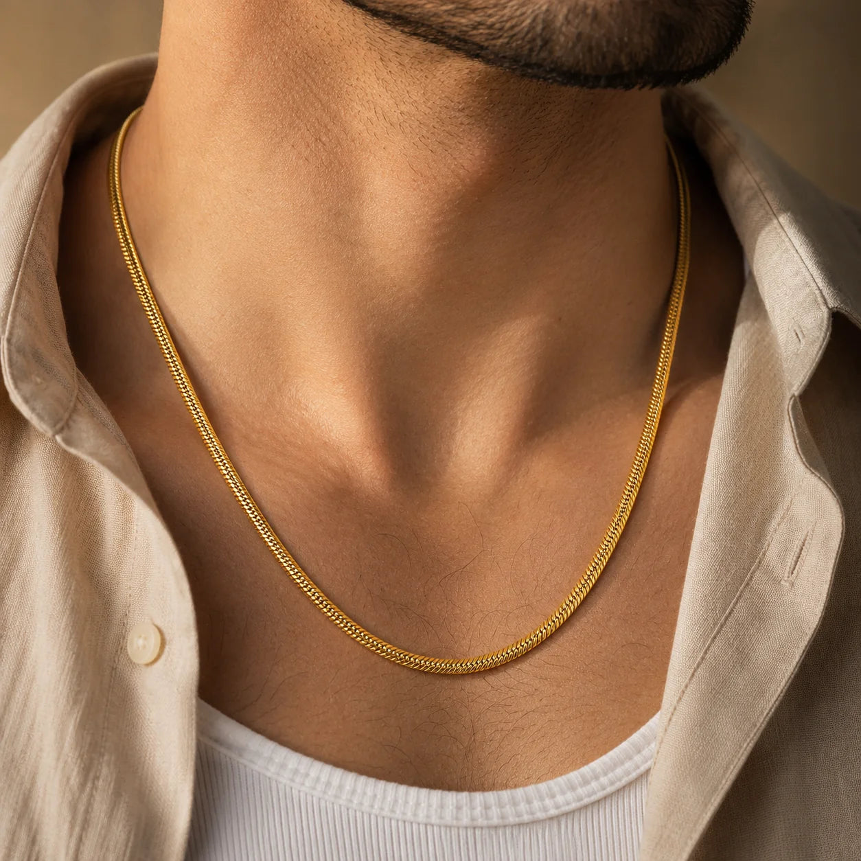 A close-up of a man's neck and chest wearing a delicate gold chain necklace over a light beige button-up shirt and white undershirt.