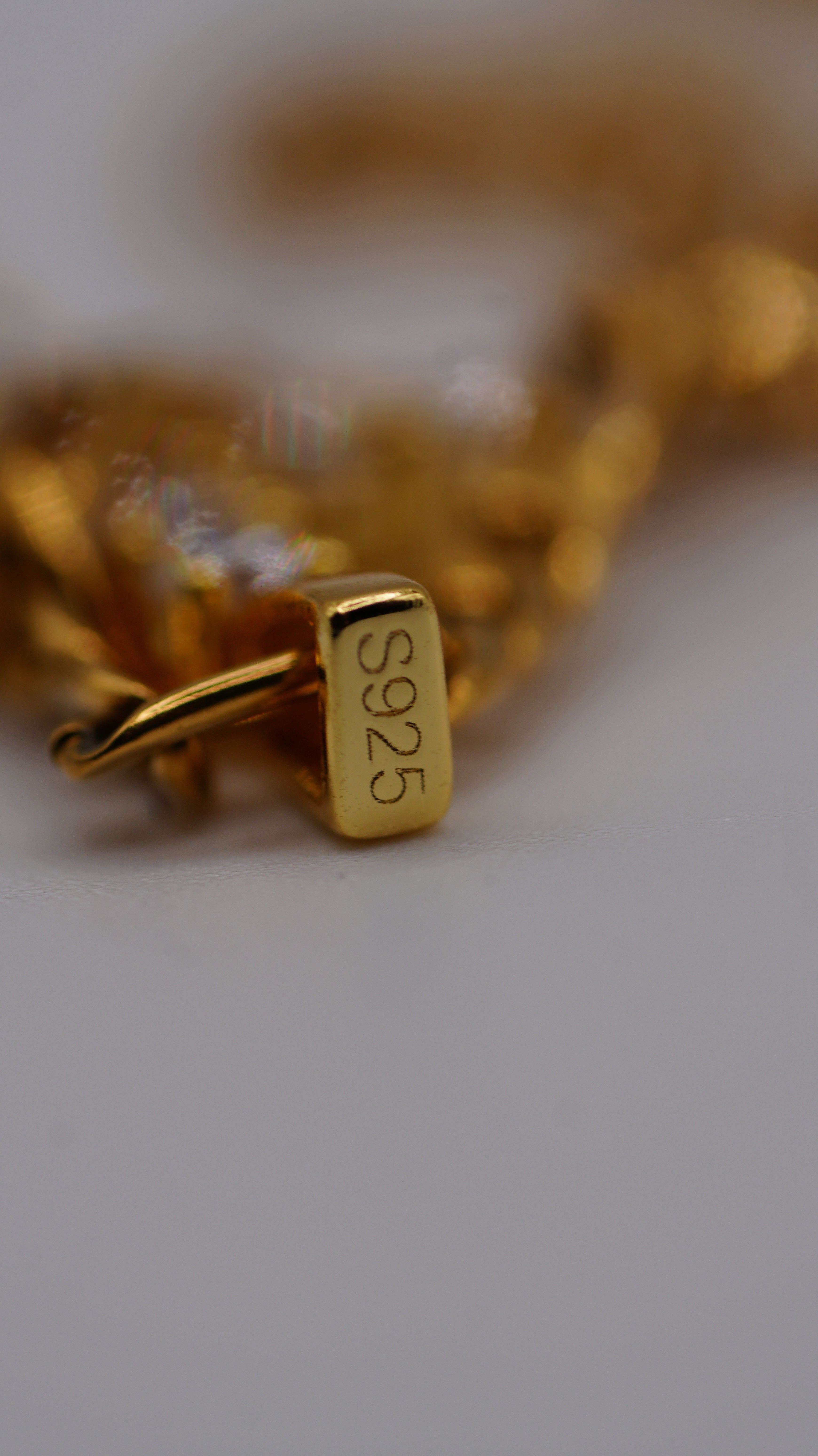 Close-up of a gold chain bracelet with a rectangular clasp engraved with 'S925', showing the sterling silver purity mark in yellow gold finish, on a light gray background.