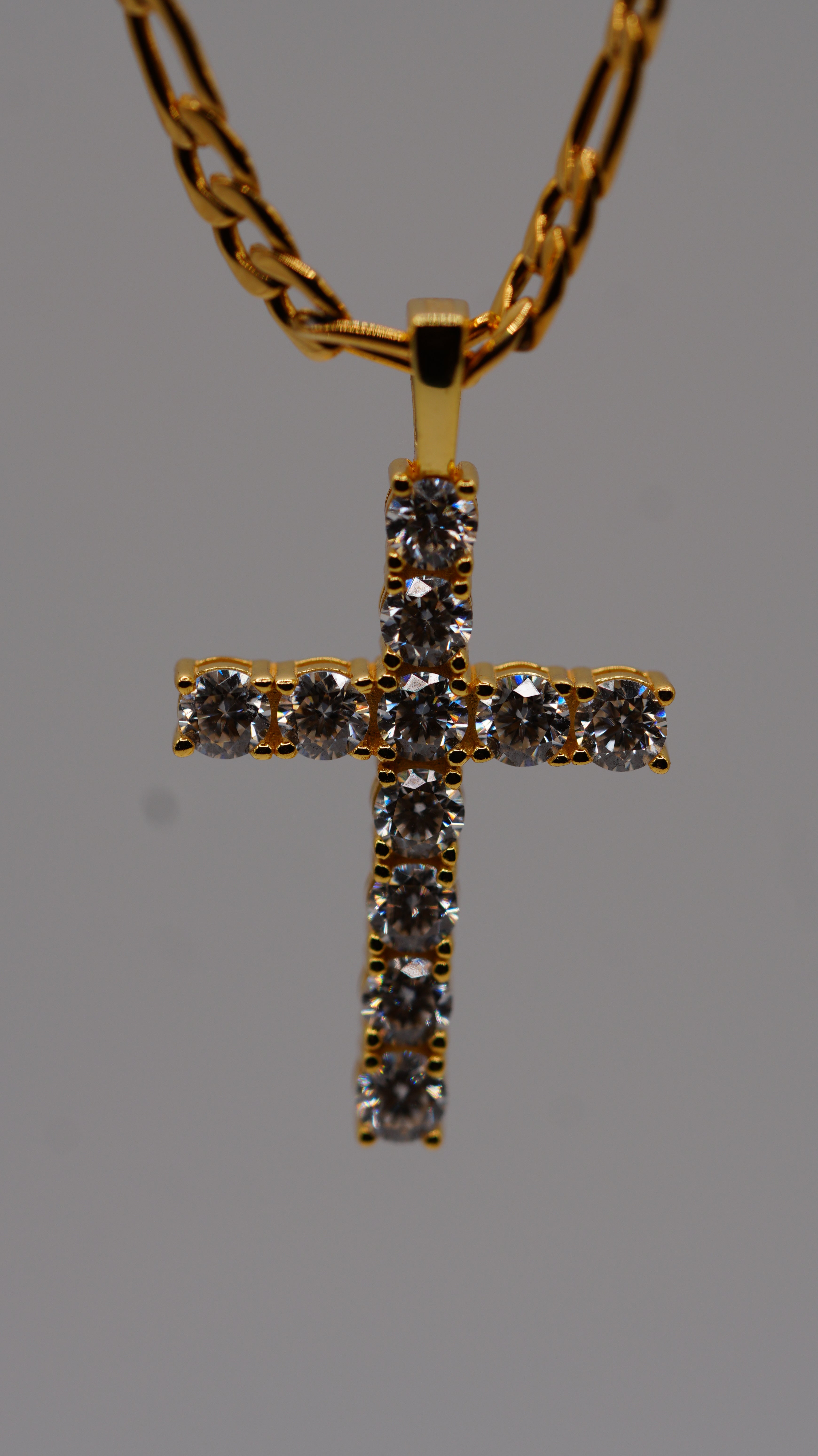 A gold cross pendant necklace adorned with clear gemstones, suspended from a gold chain against a solid gray background.