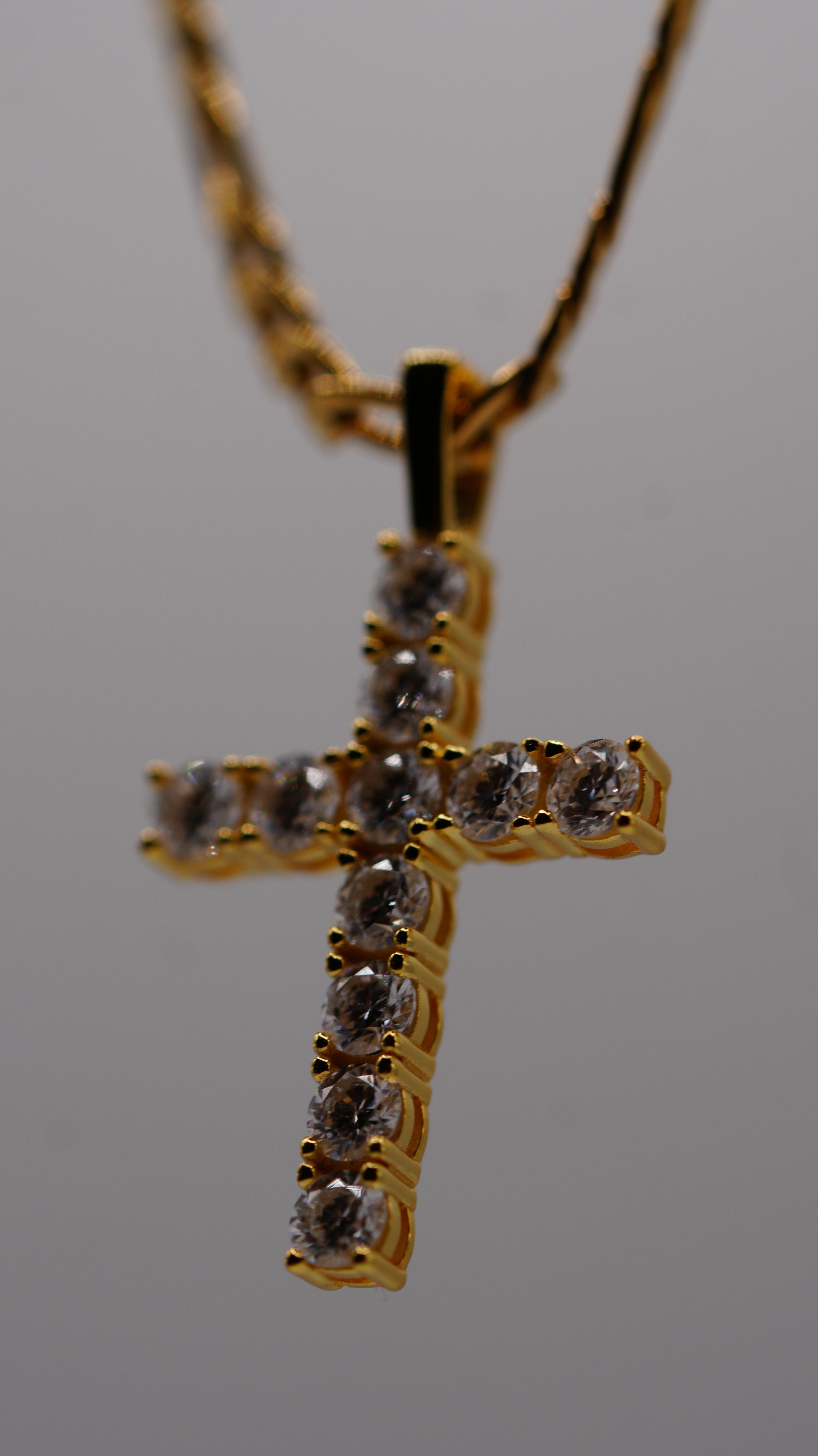 A gold cross pendant necklace adorned with clear round-cut gemstones, suspended from a gold chain against a neutral gray background.