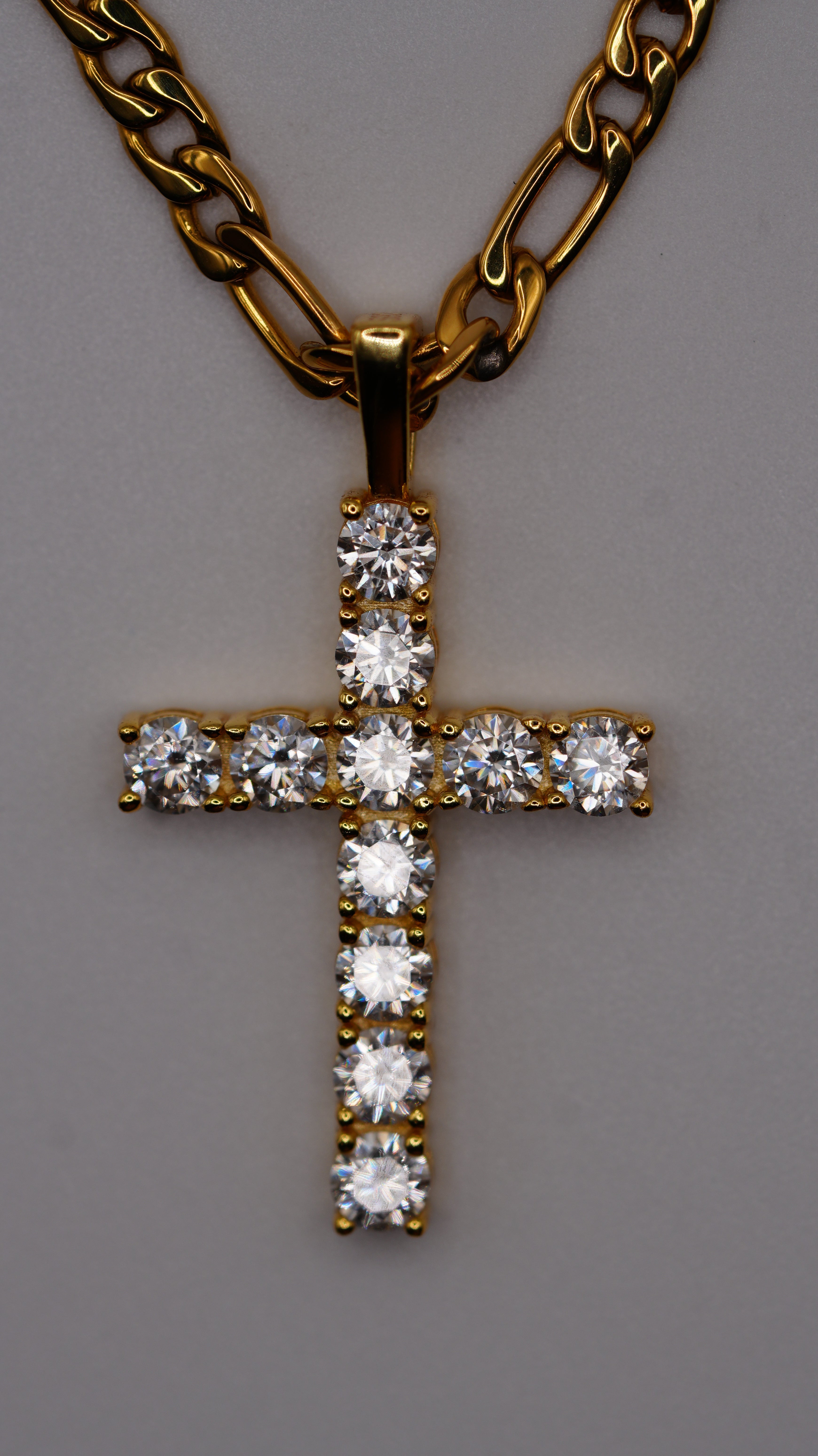 A gold cross pendant adorned with clear round-cut gemstones, shown on a plain gray background with a gold chain attached.