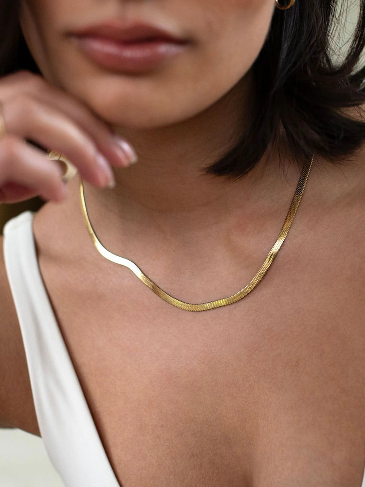 A close-up of a woman wearing a delicate, wavy gold chain necklace. The shot focuses on the jewelry, showing the textured gold chain draped around her neck and collarbone, with her hand resting near her face.