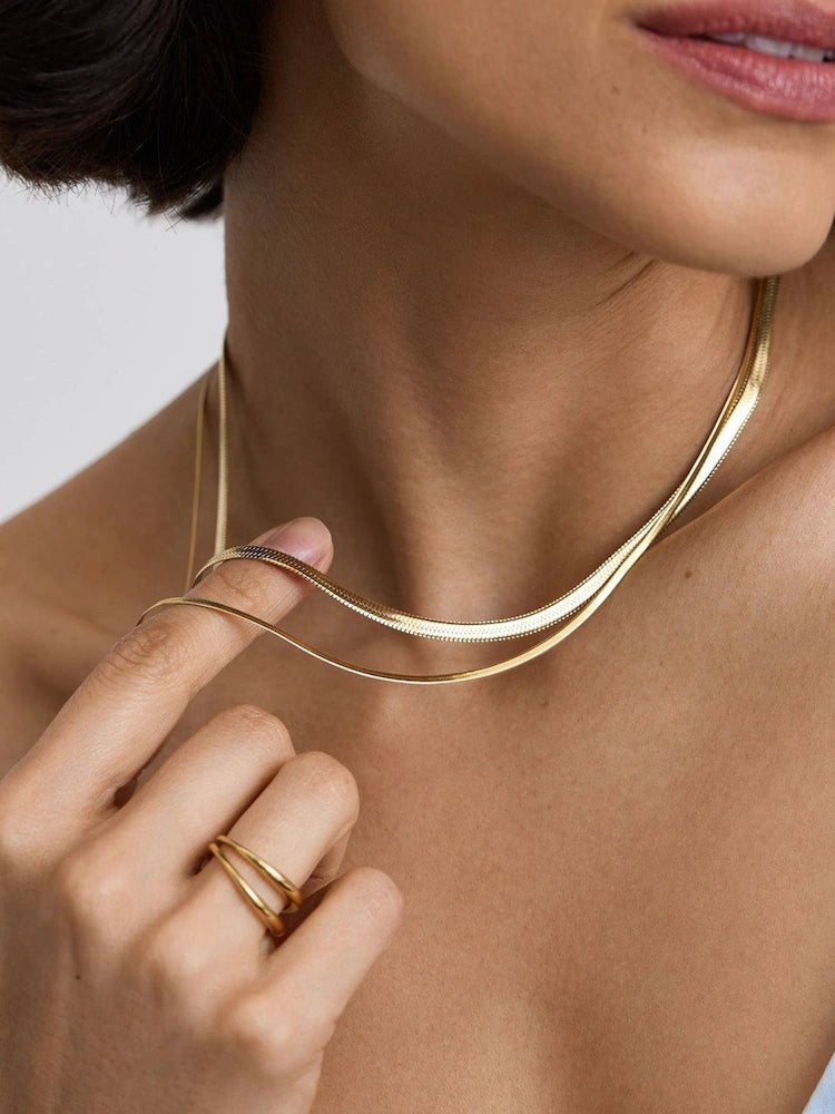 A close-up lifestyle shot of a model wearing a double-strand, twisted gold snake chain necklace and matching gold rings on her fingers.